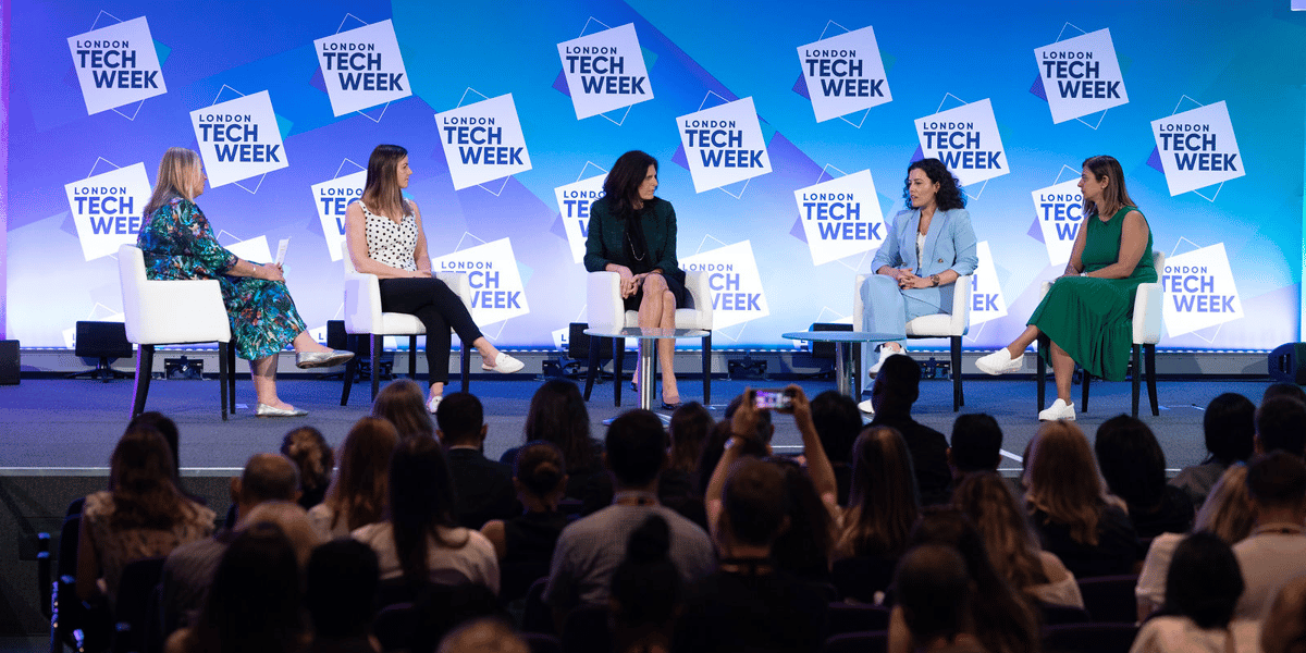Five women seated on a blue background stage in front of an audience, in discussion with one another