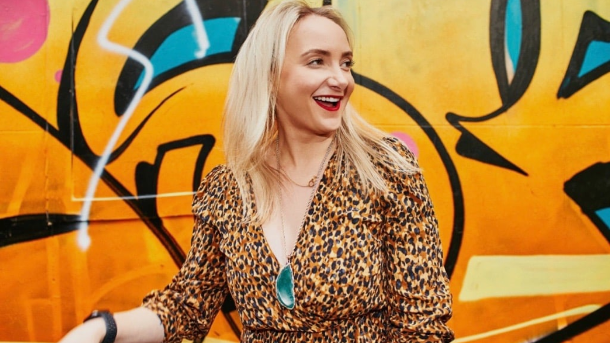 The image features Alicia Teagle smiling and looking away from the camera. Alicia is wearing a leopard print clothing, a black watch on her right wrist, and a necklace with a large green pendant. The background is a vibrant graffiti wall with various colors including yellow, blue, orange, and pink, with some abstract designs and shapes. The overall vibe is casual and joyful, with an urban, artistic setting.