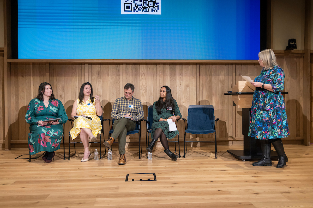 Four panellists sit at the front of a wood auditorium with a blue presentation screen behind them. A moderator stands to the right next to a lectern.